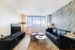 A modern living room with a flat-screen TV, a black leather sofa, two small round tables, and a patterned wall. - Elevated City Living | UWS | Doorman & W/D (New York)