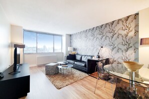 A modern living room with a black leather sofa, a glass coffee table, and a city view through large windows.