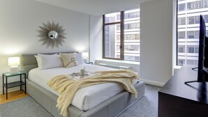 Un dormitorio moderno con una cama grande, una mesita de noche con lámpara, un espejo y vistas a edificios a través de una ventana.