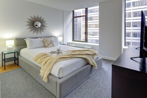 Una camera da letto moderna con un letto grande, un comodino con una lampada, uno specchio e vista sugli edifici attraverso una finestra.