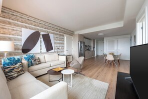 A modern living room with a sofa, coffee table, and TV. There is a kitchen in the background with a refrigerator and cabinets.