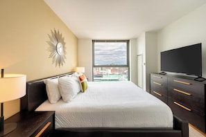 A modern hotel room with a large bed, a TV, a nightstand, and a view of the city through a large window.