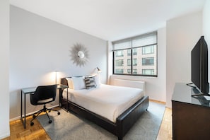 Une chambre moderne avec un lit, un bureau, une chaise et une grande fenêtre donnant sur des immeubles.