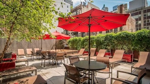 An outdoor patio with tables and chairs, red umbrellas, and a wooden fence. - Brooklyn Heights Living | Doorman, On-Site W/D (Brooklyn)