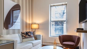 Una sala de estar moderna con un sofá blanco, un sillón marrón, una mesa de centro redonda, una lámpara y un ventanal con vistas a edificios.