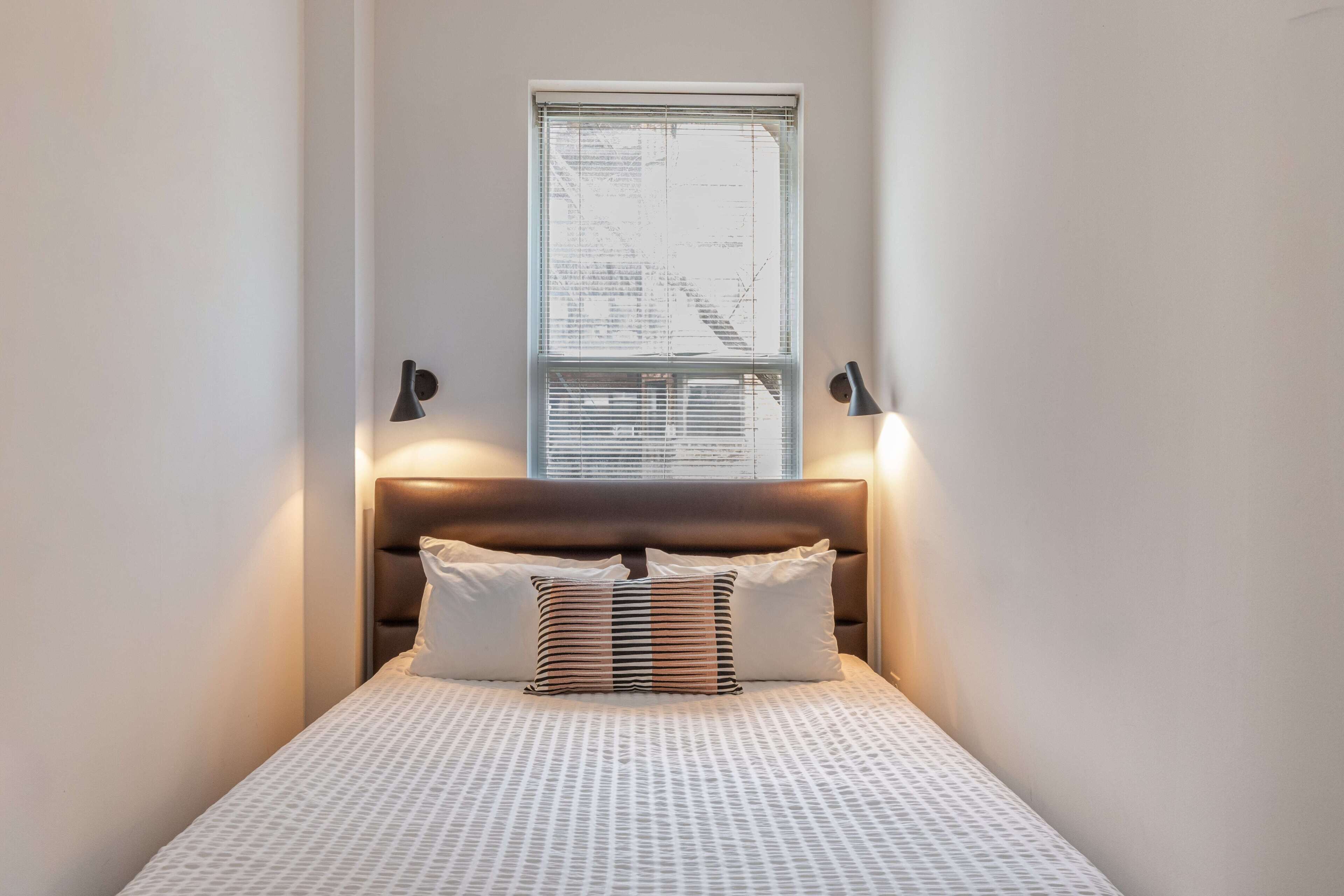 A narrow hotel room with a bed, a window with blinds, and two wall-mounted lamps.