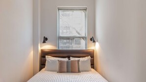 Habitación de hotel estrecha con una cama, un ventanal con persianas y dos lámparas fijadas a la pared.