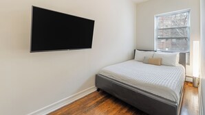 Quarto com cama, televisão montada, janela com persianas e chão de madeira.