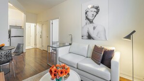 Uma sala de estar moderna com um sofá branco, uma mesa de centro de vidro com flores, um abajur preto e um grande retrato em preto e branco na parede.
