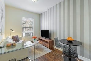 A modern living room with a glass coffee table, a flat-screen TV, a dining table with chairs, and a window with blinds.
