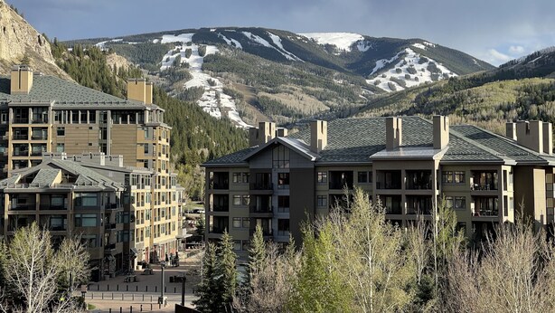 A modern building complex with multiple floors and balconies, surrounded by trees and mountains in the background.