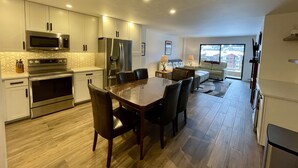 A modern kitchen with stainless steel appliances, a dining area with a wooden table and chairs, and a living room with a sofa and a large window.