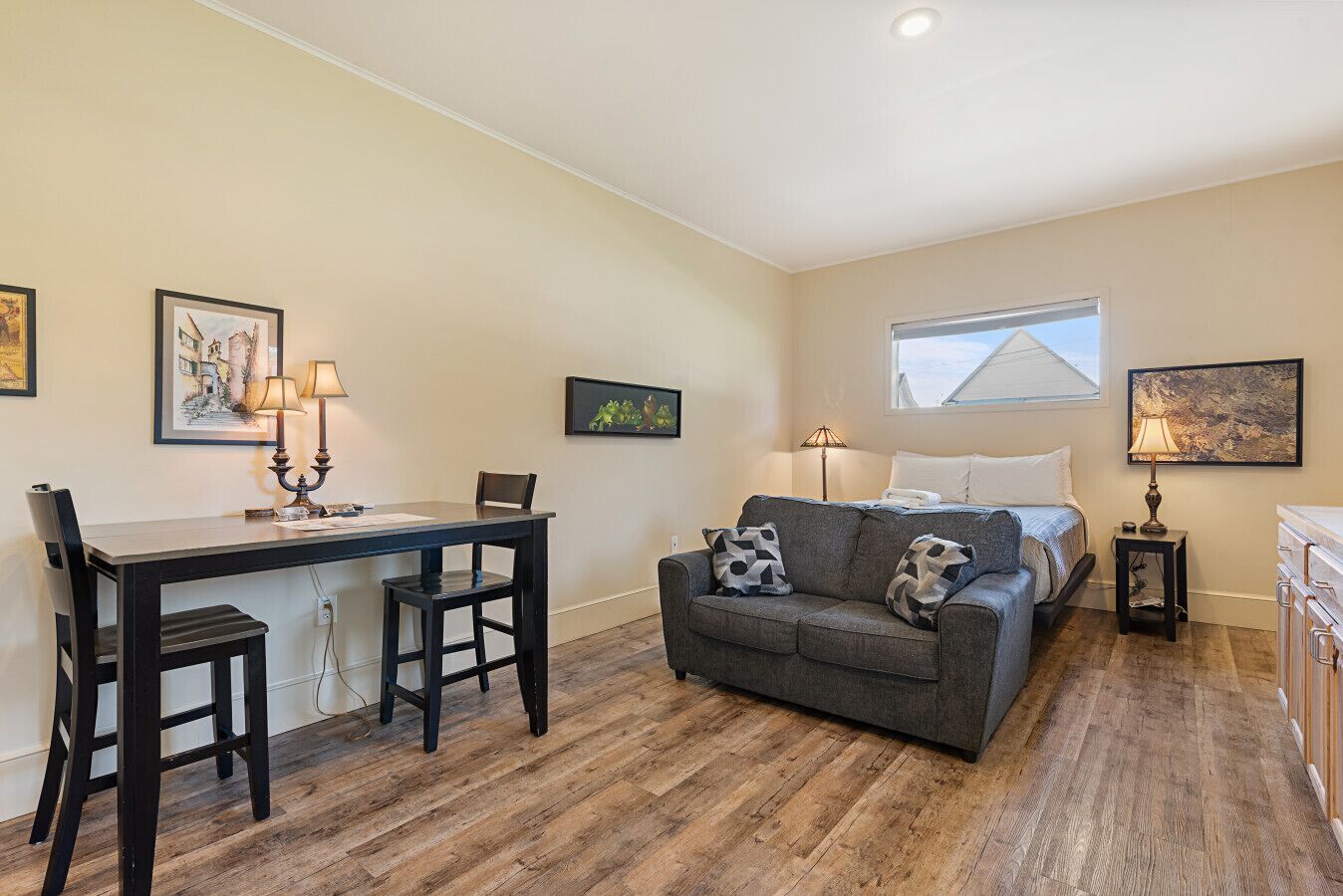 A compact living space with a dining table, two chairs, a sofa with patterned cushions, a bed, a small side table with a lamp, and a window with a view.