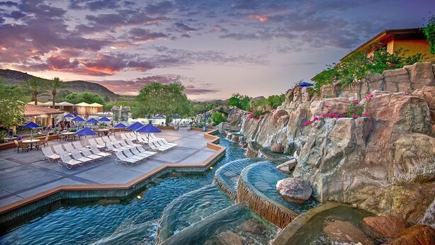 Un espace piscine aménagé dans un complexe hôtelier, avec des chaises longues et des parasols, entouré d’un paysage rocheux et illuminé par un ciel au coucher du soleil.