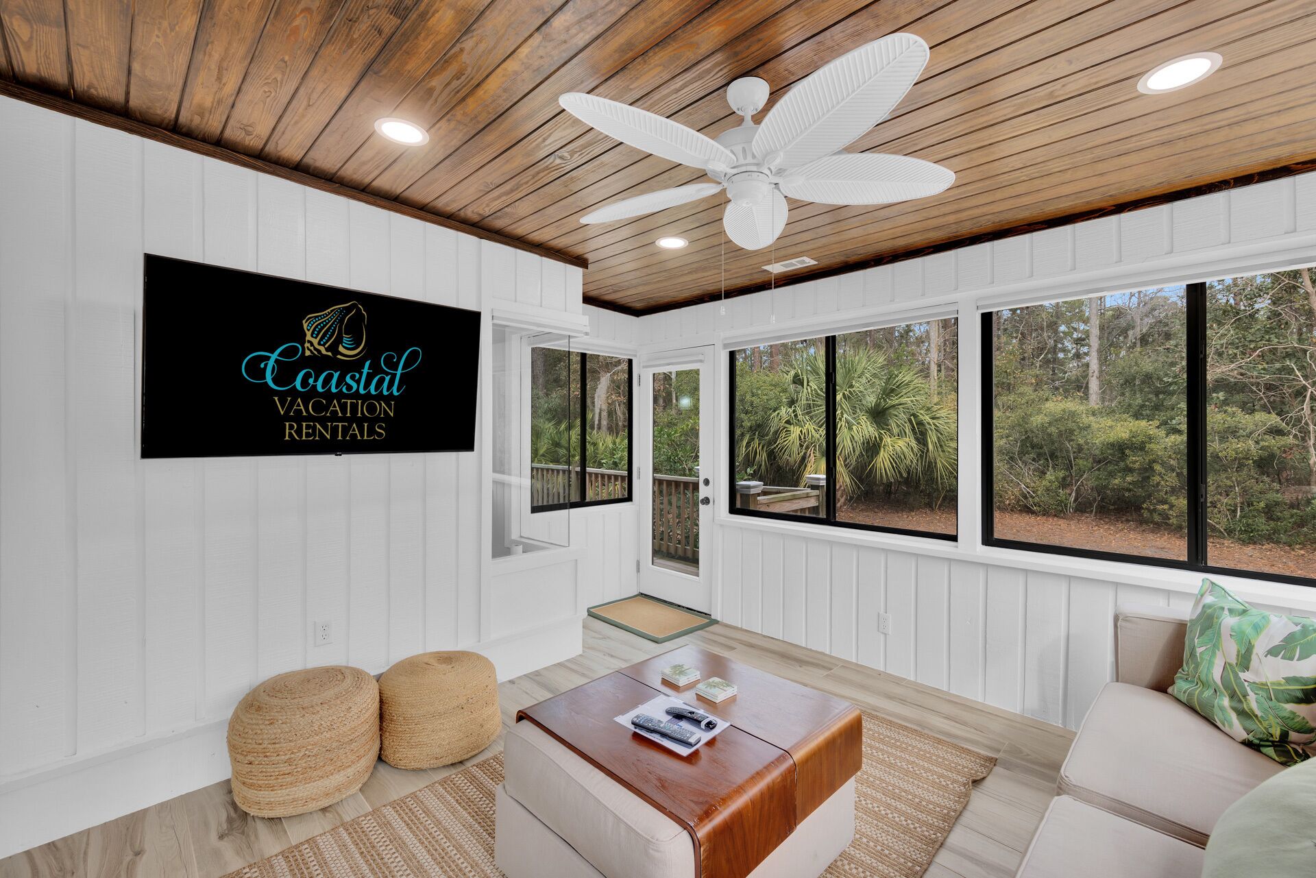 A spacious room with a ceiling fan, large windows offering a view of the outdoors, a mounted sign for Coastal Vacation Rentals, and a wooden coffee table.