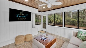 Ein geräumiges Zimmer mit Deckenventilator, großen Fenstern mit Blick ins Freie, einem an der Wand befestigten Schild für Coastal Vacation Rentals und einem Holztisch.