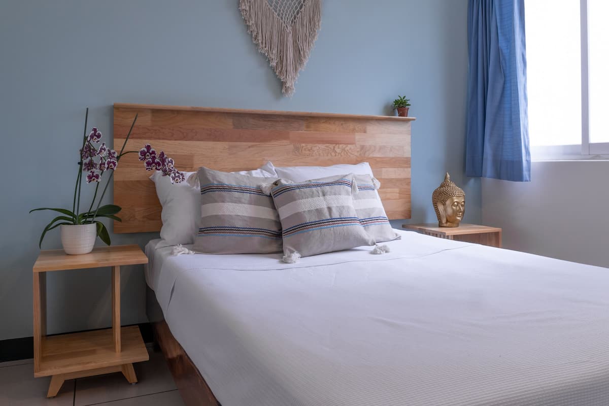 A neatly made bed with a wooden headboard, two bedside tables with decorative items, and a wall-mounted macrame piece.