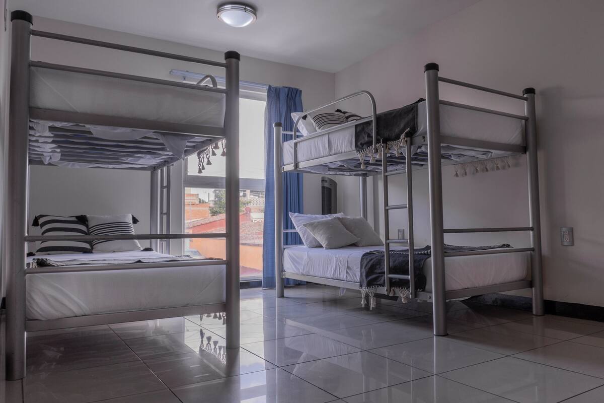 A dormitory room with bunk beds, a window with curtains, and a tiled floor.