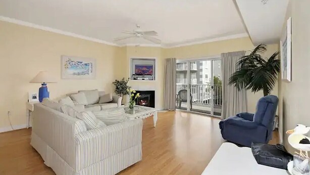 A living room with a sofa, armchair, and a coffee table. There is a ceiling fan, a fireplace, and a balcony with a view. - ⭐️Astoria 207 Ocean/Bayside Views Rooftop Pool Linens⭐️ (Ocean City)