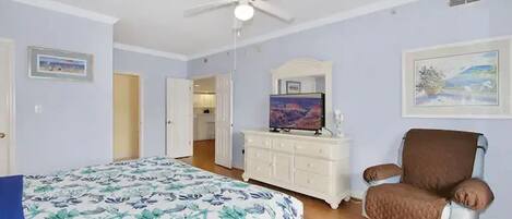 Un dormitorio con una cama, un tocador, una silla, un ventilador de techo y un televisor.