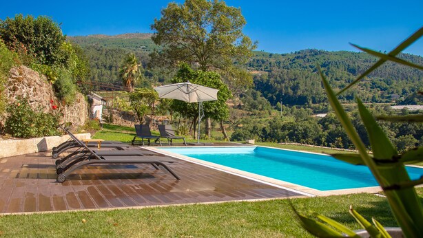 A swimming pool with a wooden deck and lounge chairs, surrounded by a grassy area and trees, with a view of hills in the background. - Buganvília Loft - Quinta do Tapadinho (Cabeceiras de Basto)