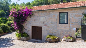 A stone building with a red-tiled roof, a wooden door, and potted plants with flowers. - Buganvília Loft - Quinta do Tapadinho (Cabeceiras de Basto)
