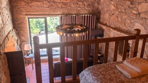 A rustic room with a wooden bed, a stone wall, a hanging light fixture, and a view of the outdoors. - Buganvília Loft - Quinta do Tapadinho (Cabeceiras de Basto)