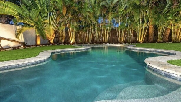 A swimming pool with a curved edge, surrounded by palm trees and a wooden fence. - Steps to Beach & Pickleball • Pool, Game Room, Putting Green & Cart (Indian Rocks Beach)