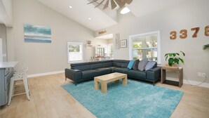 A spacious living room with a sectional sofa, a wooden coffee table, and a blue rug. There is a ceiling fan, a kitchen area in the background, and a large window with blinds. - Steps to Beach & Pickleball • Pool, Game Room, Putting Green & Cart (Indian Rocks Beach)