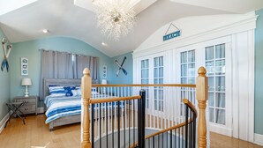 A bedroom with a bed, a nightstand, a lamp, a chandelier, a staircase, and a window with curtains. - Steps to Beach & Pickleball • Pool, Game Room, Putting Green & Cart (Indian Rocks Beach)