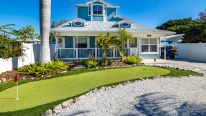 A two-story house with a covered porch, a small front yard with artificial grass, and a gravel driveway. - Steps to Beach & Pickleball • Pool, Game Room, Putting Green & Cart (Indian Rocks Beach)