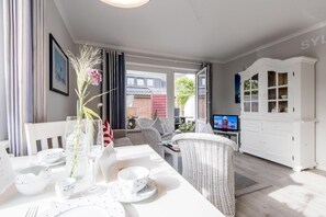 A dining area with a table set for two, a wicker chair, a cabinet, and a television in the background.