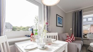 Quarto bem iluminado com uma mesa de jantar posta para dois, um sofá com almofadas listradas e um quadro na parede.