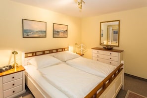 A neatly made bed with white linens, a bedside table with a lamp, a dresser with drawers, and two framed pictures on the wall.