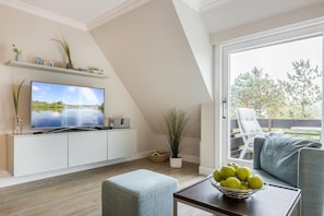 A modern living room with a TV, white cabinetry, a dining area with a table and chairs, and a balcony with outdoor furniture.