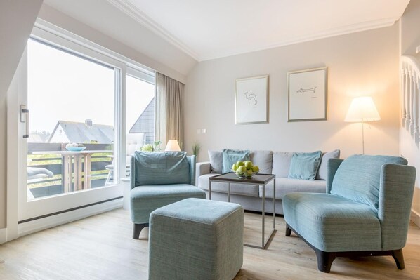 Ein modernes Wohnzimmer mit einer Couch, zwei Sesseln, einem Couchtisch und einer Lampe. An der Wand hängen zwei gerahmte Bilder und es gibt ein Fenster mit Vorhängen.