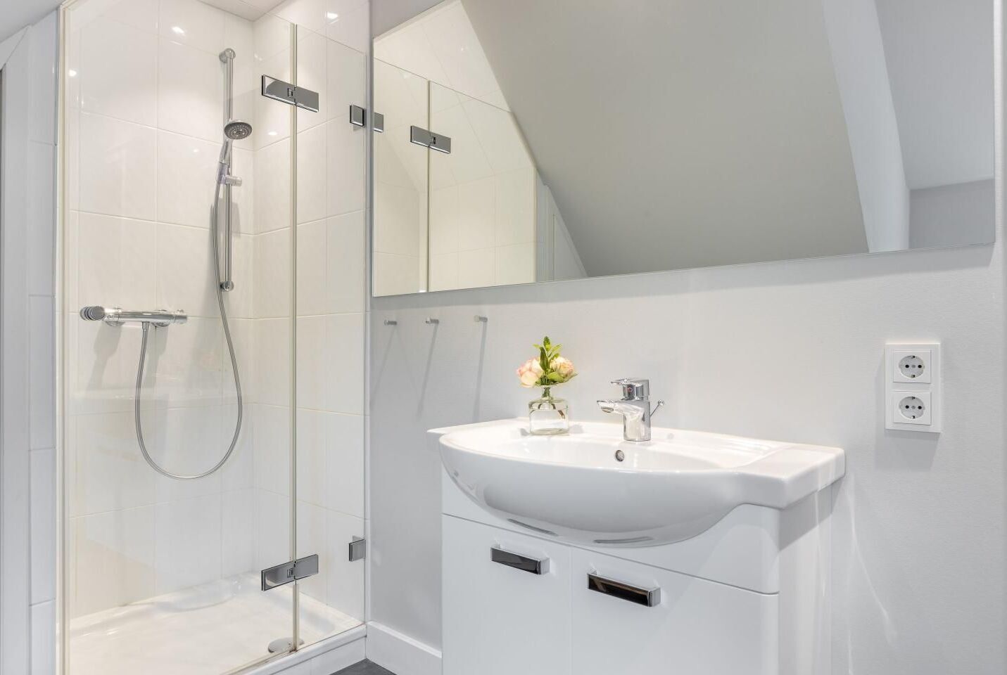 A modern bathroom with a glass-enclosed shower, a white sink with a vase of flowers, and a mirror above the sink.