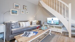 Un salon moderne avec un canapé gris, une table basse en bois, un téléviseur posé sur un meuble blanc et un escalier doté de balustrades blanches.