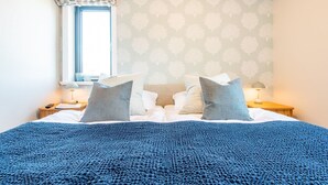 Un lit bien fait, recouvert d’une couverture bleue, avec deux oreillers et une fenêtre ornée d’un rideau à motifs.