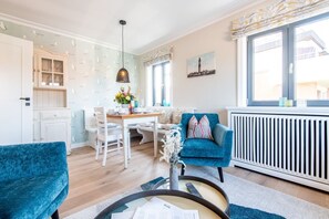A cozy living room with a dining area, a blue armchair, a white cabinet, and a window with a view of the outside. - Stadthaus Boysenstraße, Whg. 5 - Stadthaus Boysen (Sylt)