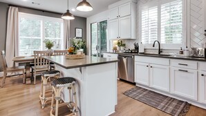 Une cuisine moderne avec des meubles blancs, un îlot central doté de tabourets, un lave-vaisselle et une grande fenêtre avec des stores.