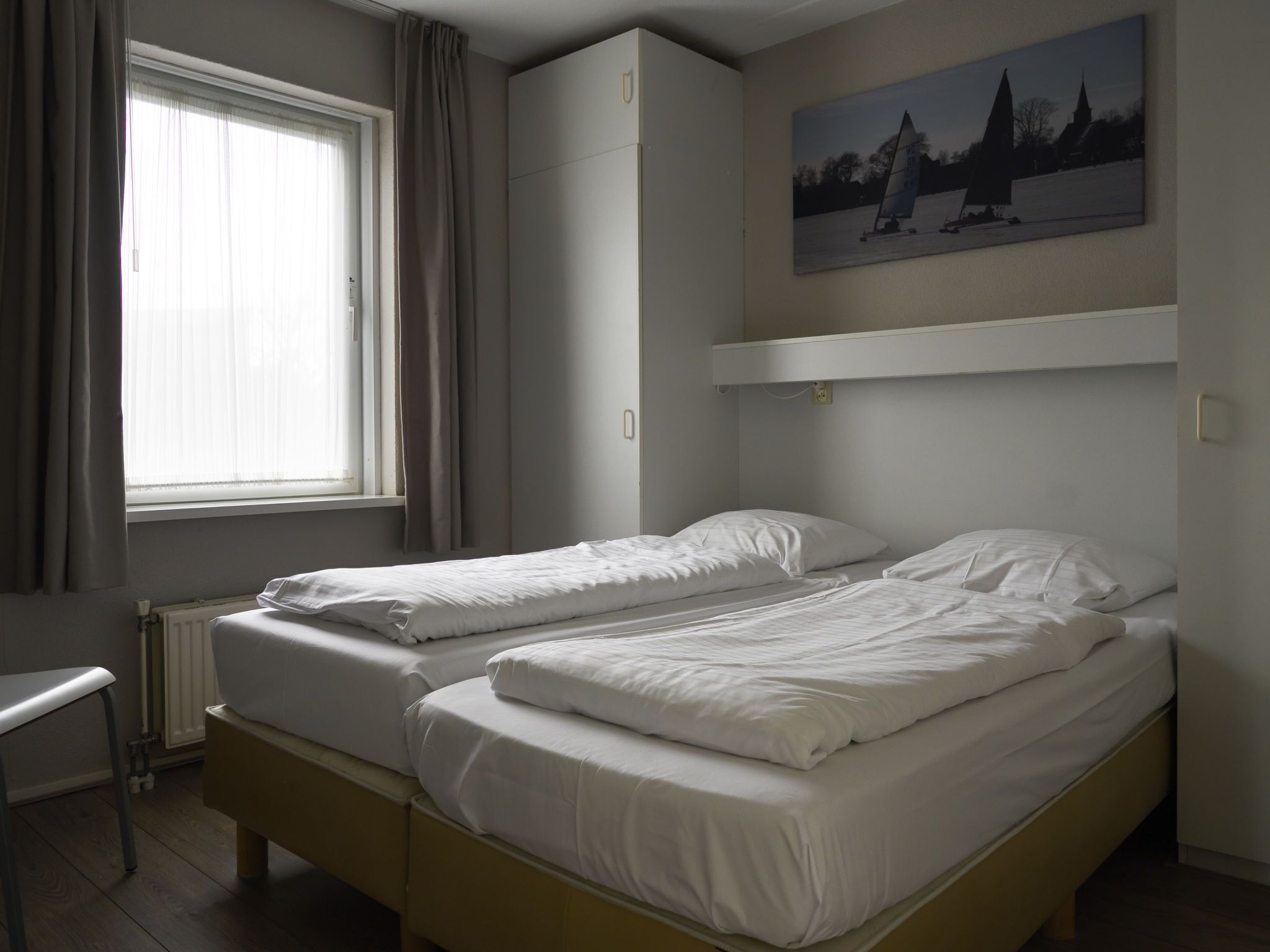 Une chambre avec deux lits simples, un radiateur, une fenêtre avec des rideaux, un tableau au mur et une armoire.