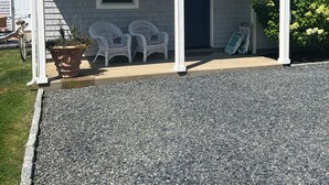 Une maison de plain-pied avec un porche couvert, une porte d’entrée et deux chaises en plastique blanches.