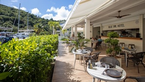An outdoor dining area with tables and chairs, a sign for the Blue Sail Hotel, and boats in the background. - Blue Sail Hotel SXM (Anse Marcel)