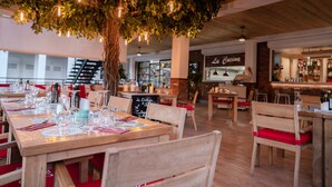 A restaurant with wooden tables and chairs, a large tree with hanging lights, and a visible menu board. - Blue Sail Hotel SXM (Anse Marcel)