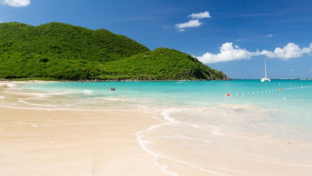 A beach with clear turquoise water, a sailboat in the distance, and lush green hills in the background. - Blue Sail Hotel SXM (Anse Marcel)
