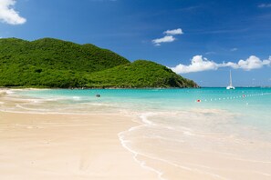 Una playa con agua turquesa cristalina, un velero a lo lejos y colinas verdes exuberantes al fondo.