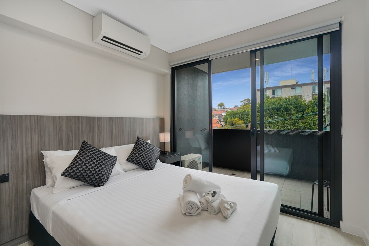 A modern hotel room with a large bed, two pillows, a rolled towel, a nightstand with a lamp, and a sliding glass door leading to a balcony.