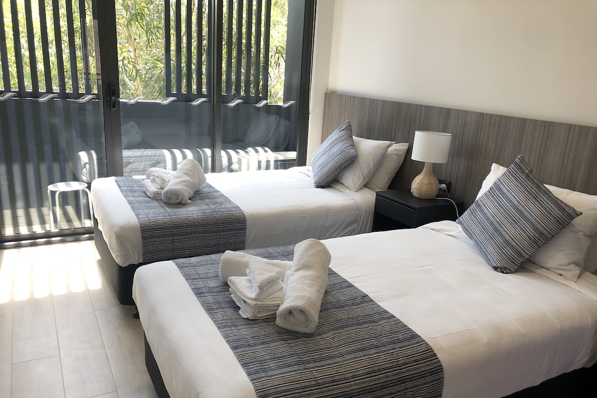 A hotel room with two beds, each with a striped bedspread and pillows, a nightstand with a lamp, and a view of a balcony with chairs.