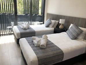 A hotel room with two beds, each with a striped bedspread and pillows, a nightstand with a lamp, and a view of a balcony with chairs. - Coogee Studio Apartments (Coogee)